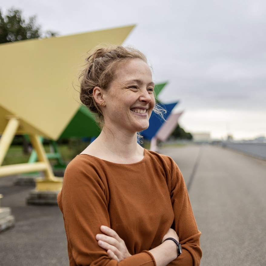 Charlotte staat lachend op een kade in de wind voor een kunstwerk van metalen bootjes, zoals je ze zou vouwen van papier