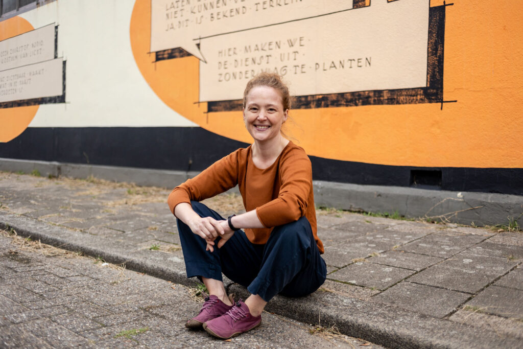 Charlotte zit op stoeprand voor een oranje graffitimuur met grote tekstballonnen. Tekst is niet leesbaar