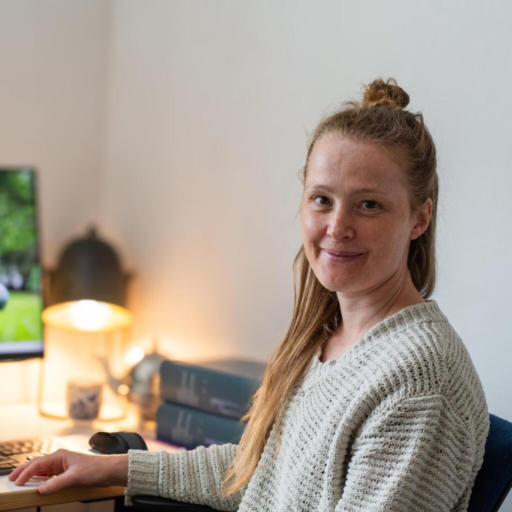 Charlotte zit aan haar bureau achter de computer. Op het bureau liggen dikke boeken en staat een theepot met een kopje