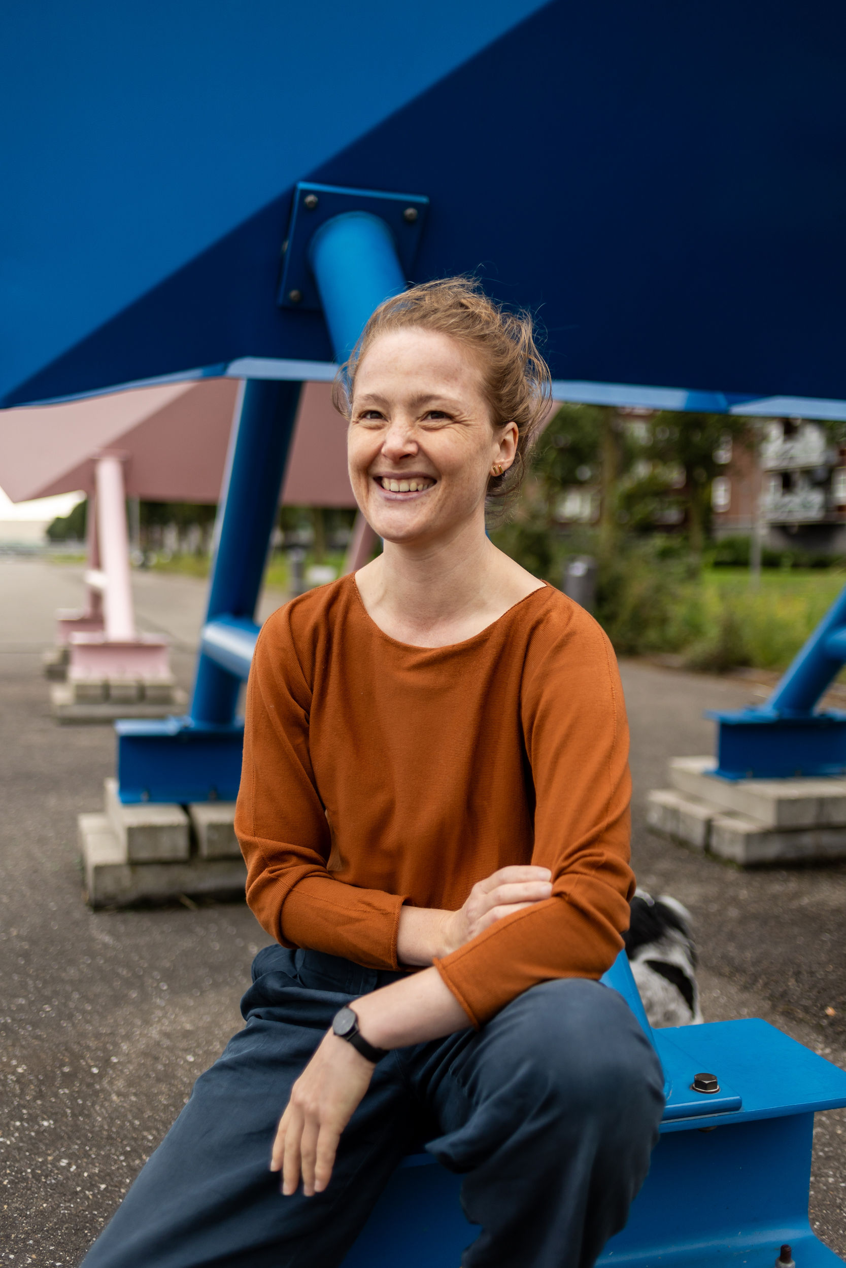 Charlotte heeft roodblond, opgestoken haar en draagt een bruine trui met boothals en een blauwe broek. Achter haar is nog net een klein, zwart-wit hondje zichtbaar