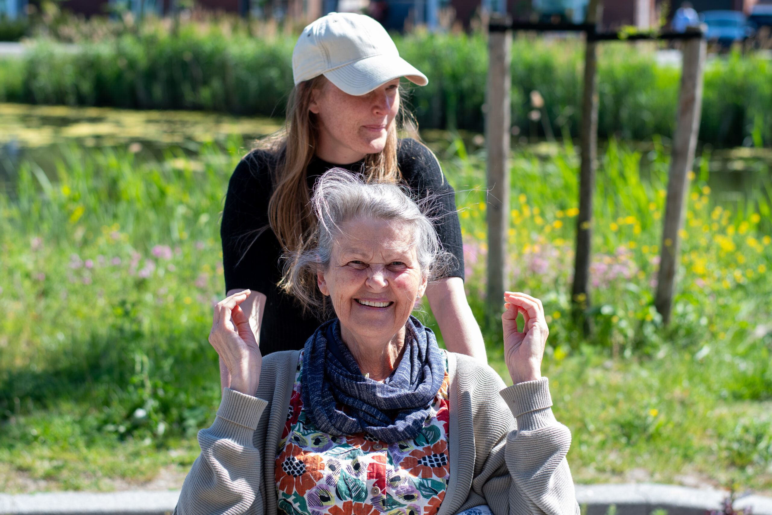 Charlotte met oudere vrouw buiten, omringd door frisgroen lentegras en wilde bloemen. Vrouw kijkt stralend in de camera