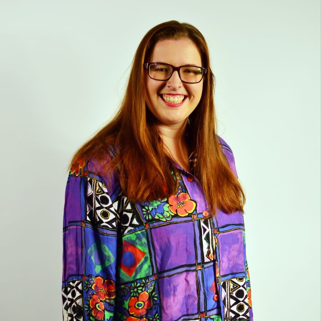 Sacha heeft lang rood haar. Ze draagt een zwarte bril met zwaar montuur, rode lippenstift en een opvallend overhemd met paars, zwart-witte patronen en bloemen erop