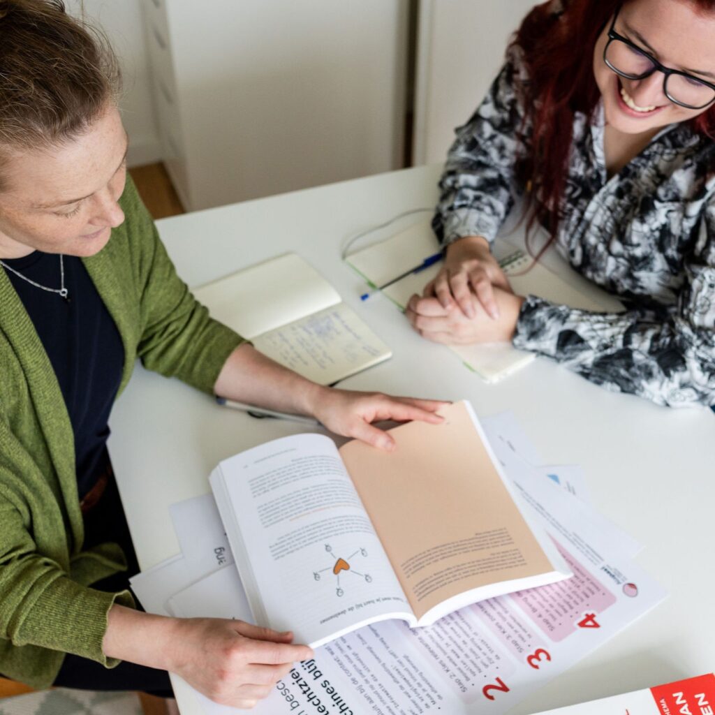 Charlotte en Sacha zitten kijken in een boek. Op tafel liggen nottieblokjes en de stappenplannen toegankelijke content