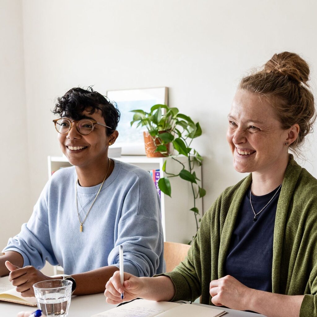 Charlotte zit aan tafel met een vrouw. Ze lachen beiden naar iemand buiten beeld. Op tafel notitieblokjes en pennen