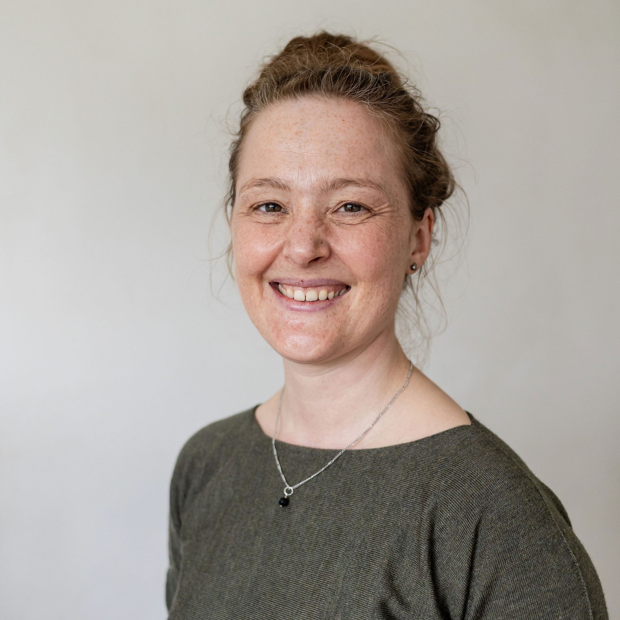 Charlotte heeft rood haar in een knot, een groene trui aan en draagt een zilveren ketting en oorbellen. Ze lacht naar de camera