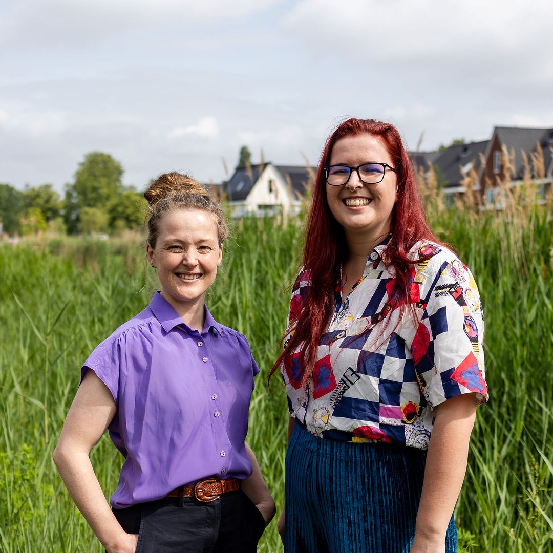 Charlotte en Sacha staan naast elkaar en lachen naar de camera. Ze staan buiten in het groen