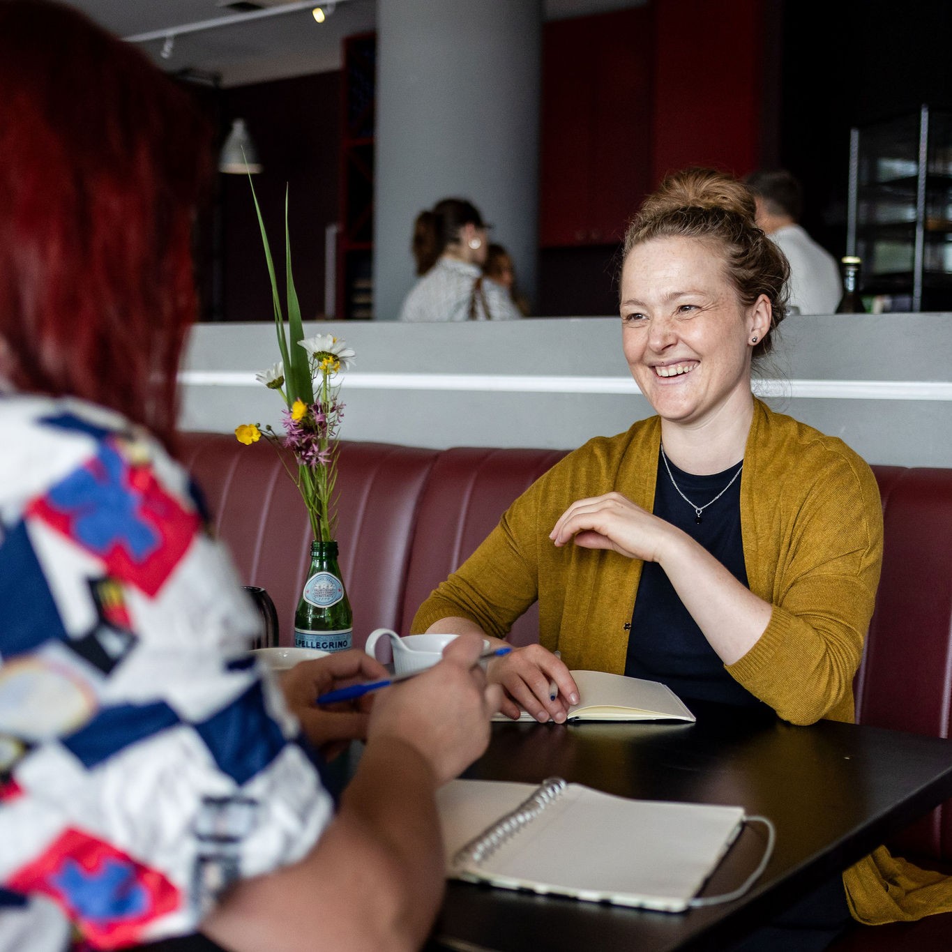 Charlotte zit tegenover Sacha aan tafel in een café. Charlotte lacht breed. Sacha heeft een pen vast. Op tafel liggen notitieboekjes