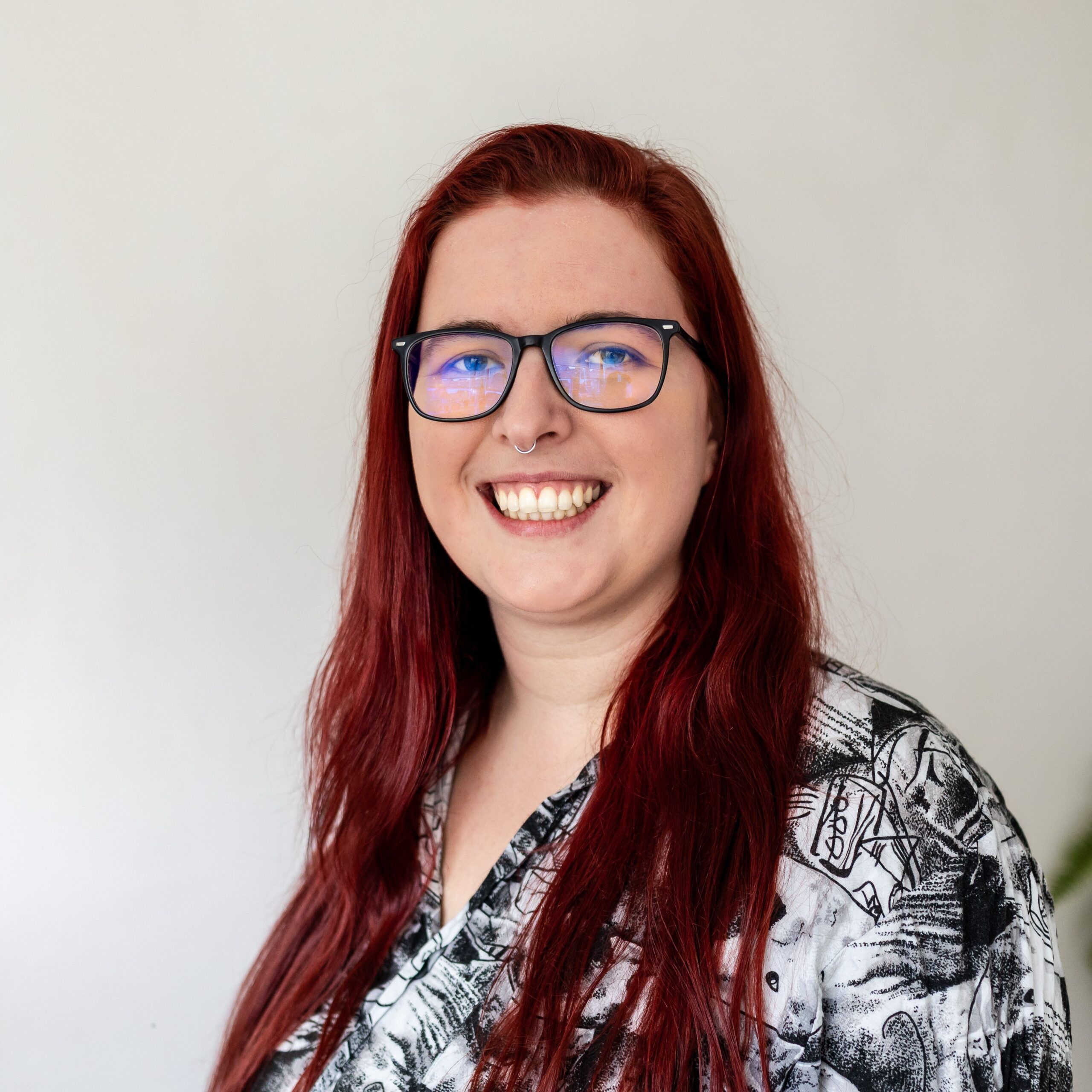 Sacha heeft rood haar, een zwarte bril met zwaar montuur en een neuspiercing. Ze draagt een zwart-witte blouse met print en lacht naar de camera